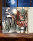 A 3D pop-up cardboard advent card featuring a festive Christmas village scene with Santa Claus in a sleigh, snow-covered trees, and a cozy house, displayed on a wooden table in a warmly lit holiday room.